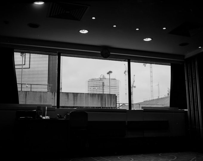 View from inside a conference room looking out through large windows at a grey urban skyline with a cylindrical tower block and construction cranes