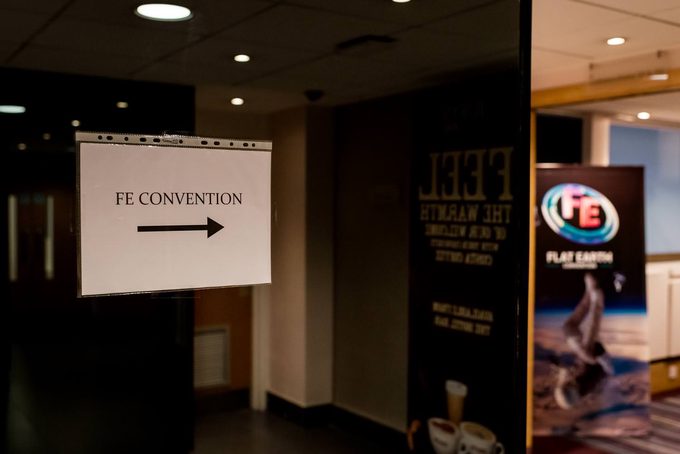 A hotel corridor with a directional sign reading ‘FE Convention’ and a Flat Earth convention banner visible through a glass door