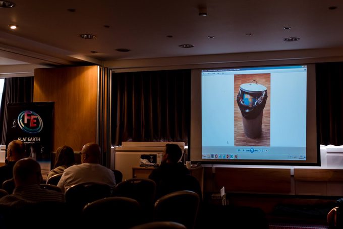 A conference room with a Flat Earth Convention banner, audience watching a projected slide showing an image of a rubbish bin