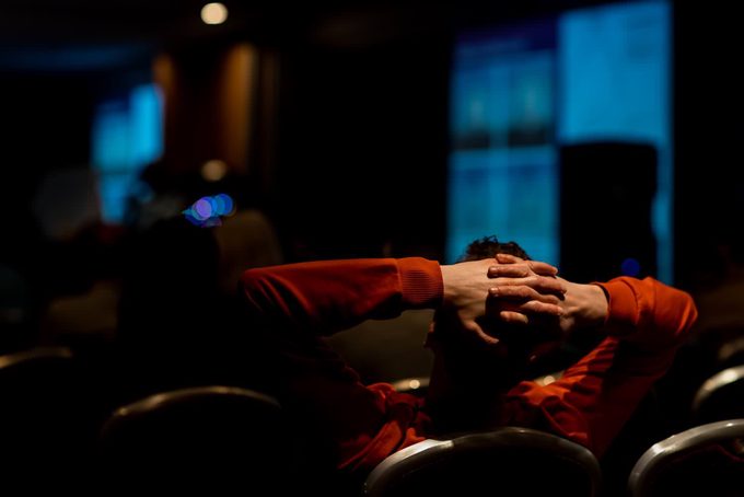 A conference attendee in a red sweater reclining with hands clasped behind their head while watching a presentation in a darkened hall