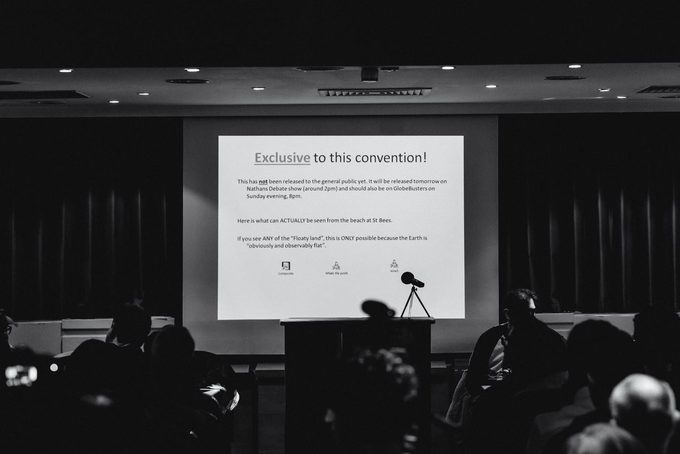 A darkened conference room with an audience watching a projected slide reading ‘Exclusive to this convention!’ claiming photographic evidence that the Earth is flat