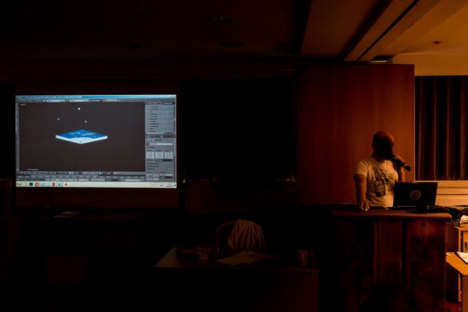 A speaker at a podium presenting to a darkened conference room, with a projected screen showing a 3D model of a flat disc representing the Earth