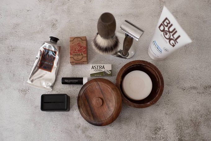 Flat lay of wet shaving accessories including L’Occitane Cade aftershave, Osma alum block, shaving brush, safety razor, Astra blades, Taylor of Old Bond Street shaving cream, wooden soap bowl, and Bulldog face wash