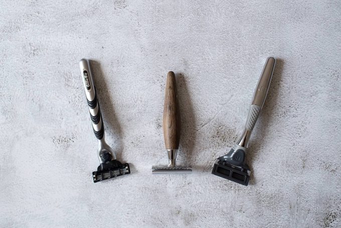 Three razors laid flat on concrete: a Gillette cartridge razor (left), a safety razor with wooden handle (centre), and a Harry’s cartridge razor (right)