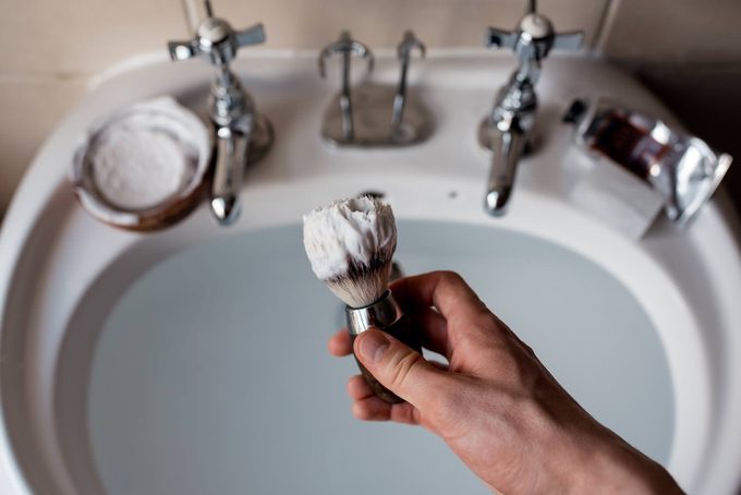 A hand holding a lather-loaded shaving brush over a bathroom sink, with soap bowl and aftershave visible