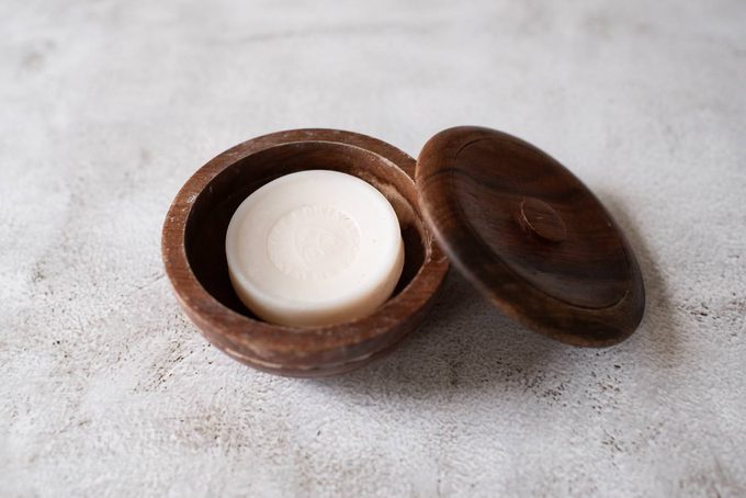 A wooden shaving soap bowl with its lid ajar, containing a puck of white shaving soap