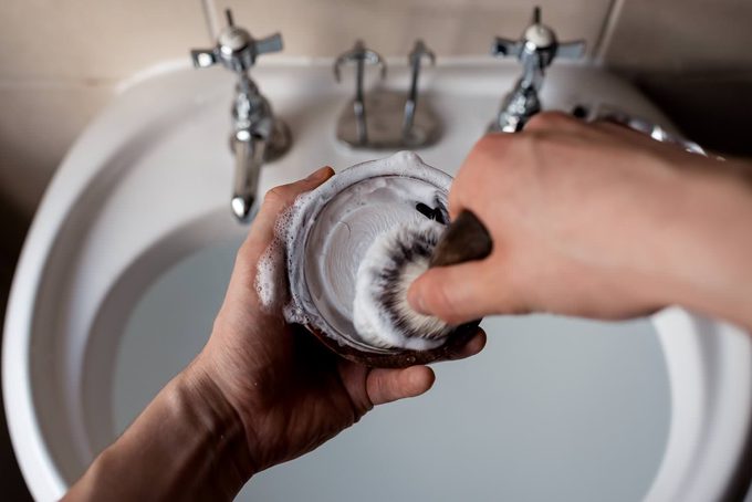 Hands loading a shaving brush with soap from a wooden bowl, building up a lather over a bathroom sink